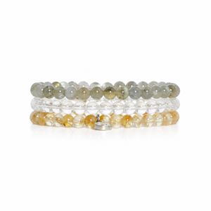 Three beaded stretch bracelets stacked together on a white background. The top bracelet features gray and olive-green beads, the middle is clear, and the bottom is a warm amber-yellow color.