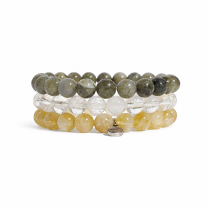 Three stacked beaded bracelets on a white background. The top bracelet features olive green and gray stone beads, the middle bracelet has clear faceted beads, and the bottom bracelet consists of translucent yellow beads with a small silver charm.