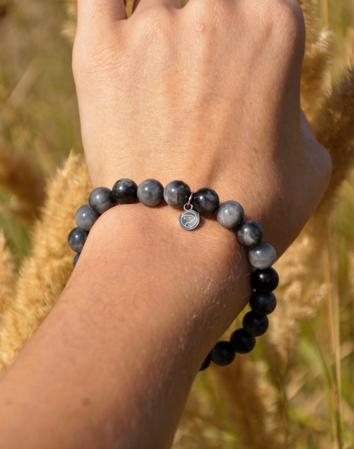 A close-up of a person's wrist wearing a beaded bracelet made of dark gray and black round beads with a small silver charm, set against a blurred natural background of dry grass.