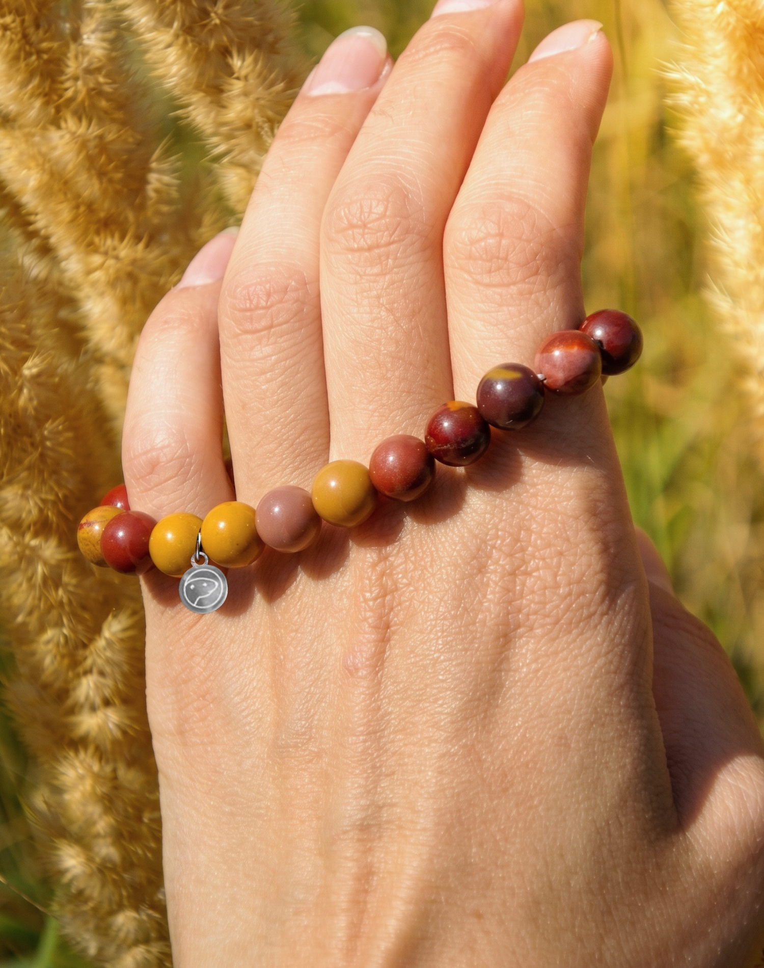 A close-up of a hand wearing a beaded stretch bracelet made of marbled brown, red, and ochre-yellow beads, with a small silver charm, set against a blurred background of golden grass.