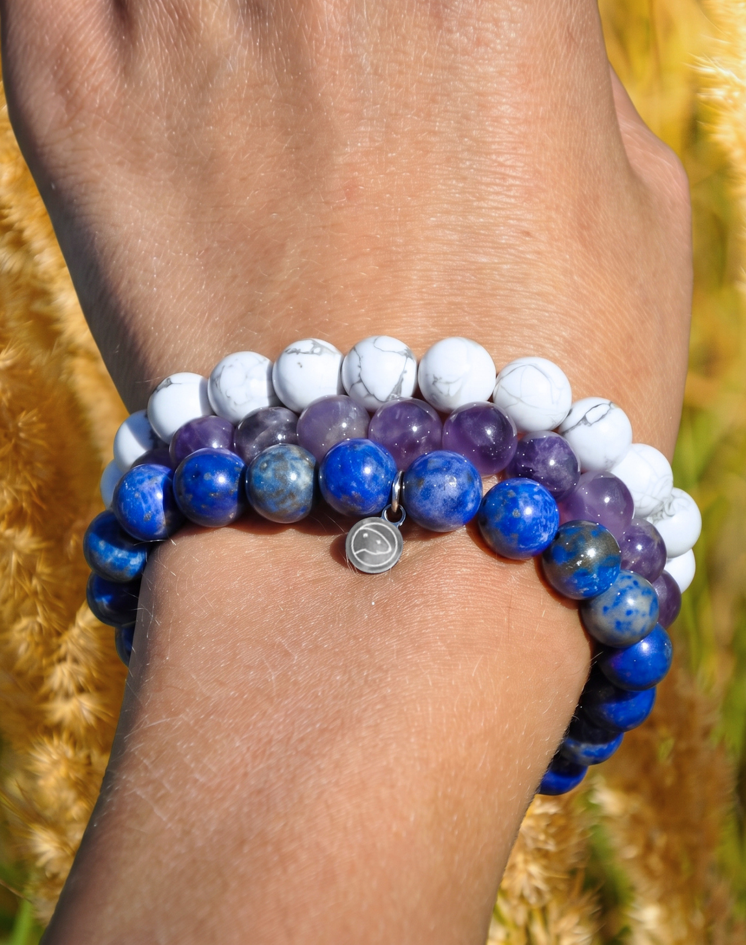 A close-up of a person's wrist wearing two beaded stretch bracelets outdoors. One bracelet is made of white marbled beads, and the other is made of deep blue and medium blue polished beads, with a small silver charm.