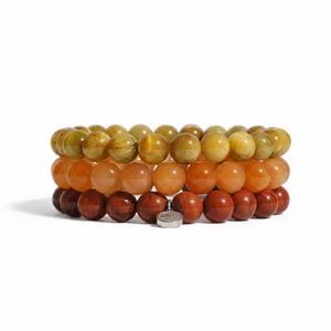 Three stacked beaded bracelets made of polished round stones in yellow-brown, orange, and reddish-brown tones, with a small silver charm at the front, photographed on a clean white background.