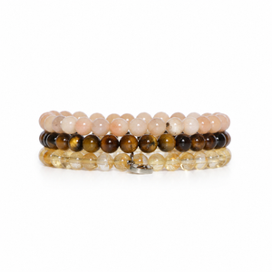 A stack of four beaded bracelets on a white background. The top bracelet features pale peach-pink beads, the second is dark brown and black tiger's eye, the third is clear yellow quartz, and the bottom bracelet has a small silver clasp.
