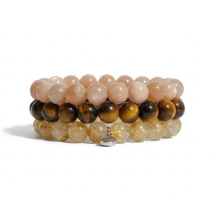 Three stacked beaded bracelets on a white background. The top bracelet is made of light peach-pink beads, the middle is made of dark brown and black tiger's eye beads, and the bottom is made of golden-brown and milky clear beads with a small silver charm.