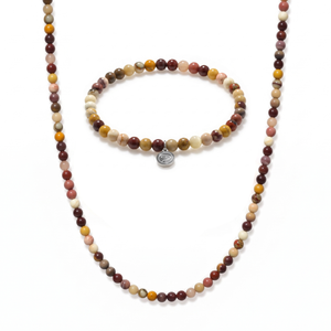 A beaded necklace and matching bracelet set on a white background. The jewelry features small round beads in earthy, multicolored shades of brown, rust red, olive green, yellow, and cream, with a small silver-toned oval charm on the bracelet.