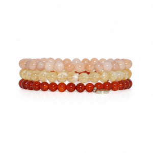 A stack of four beaded stretch bracelets on a white background. The top bracelet is made of pale peach-pink beads, the second is translucent light yellow beads, the third is opaque dark red beads, and the bottom is a small silver-toned charm with a clear stone.